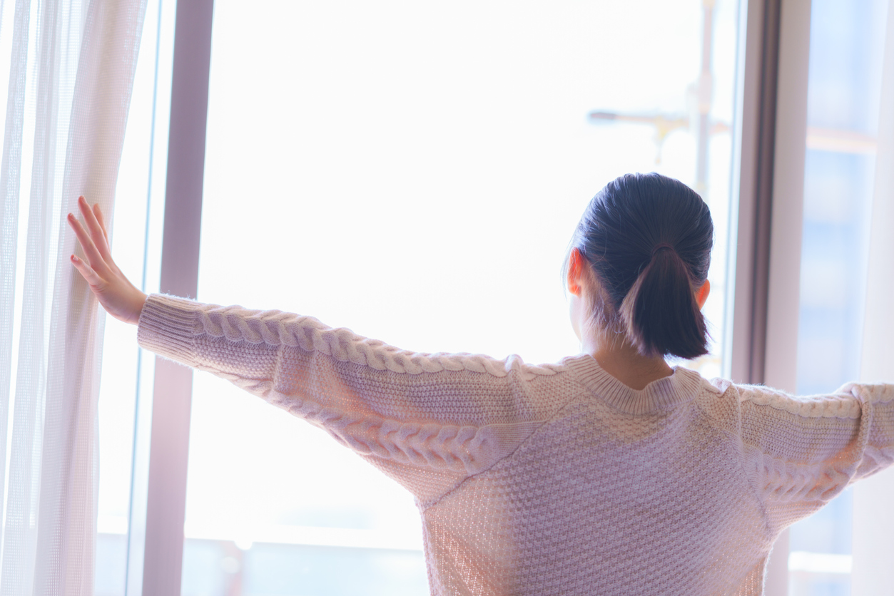 Asian woman, who is wearing a white sweater, opens curtains of a window on a clear day.